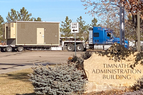 Photograph of a large truck carrying a fiber hut. The truck sits behind the Timnath Administration Building monument sign.