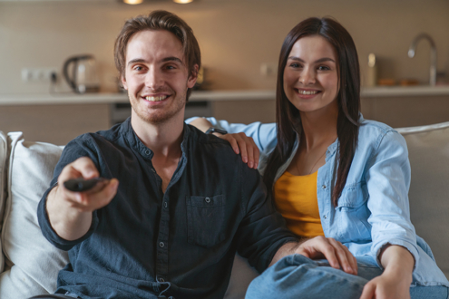 Photograph of a man and woman sitting on a couch. The man is pointing a TV remote control.