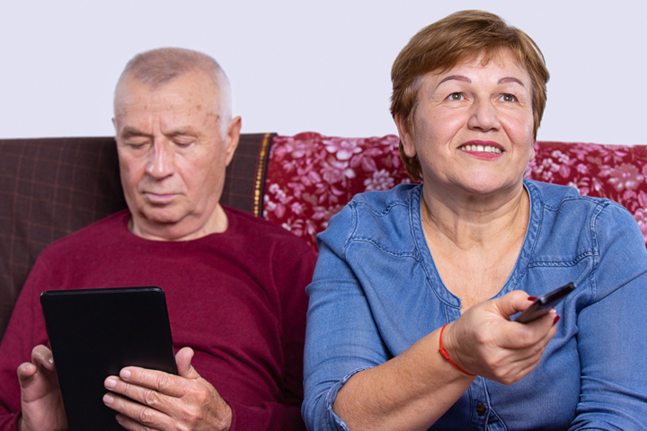 Senior married couple choosing movie on tv. Online broadcast from digital tablet on smart television. Elderly people at home relaxing on couch Photograph of two adults sitting on a couch. One is looking at a tablet and the other is pointing a remote control.